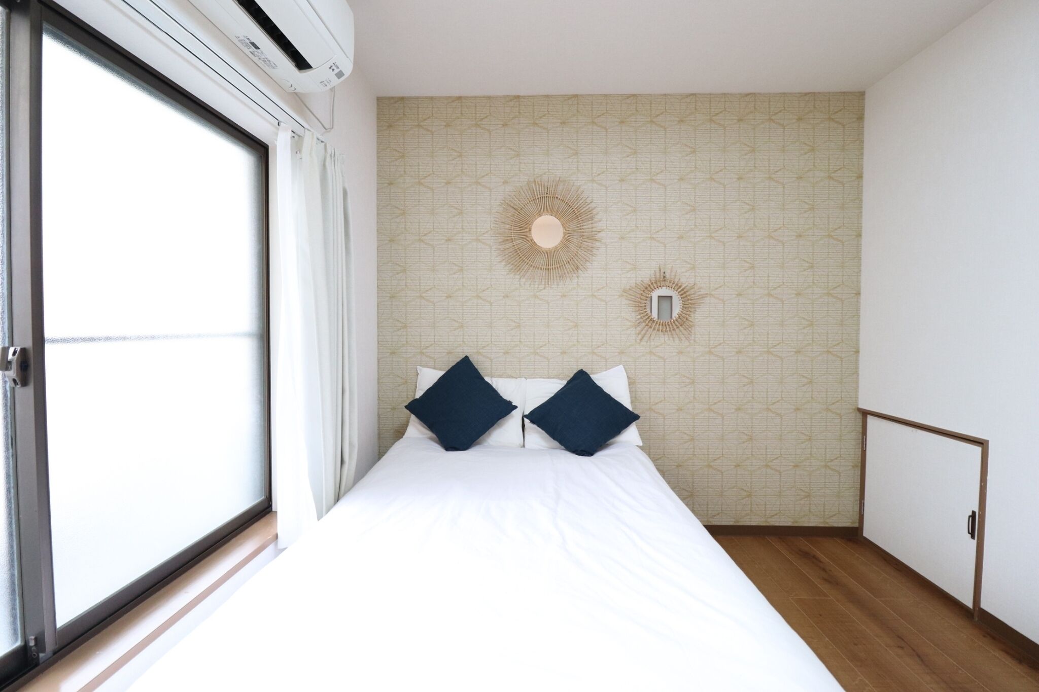 A bedroom with a large window, a bed with white bedding and two dark pillows, a geometric patterned wall, and a small wall-mounted light fixture.