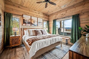 A wooden cabin bedroom with a bed, a nightstand, a bench, and a view of the outdoors. - Mountain Views, Barrel Sauna, Hot Tub, 3 King Suites, Game Room, Pool (Sevierville)
