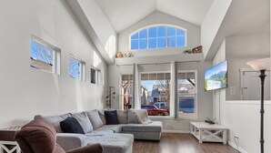 A spacious living room with a large sectional sofa, a flat-screen TV mounted on the wall, and a high ceiling with skylights. - Spacious Modern Home—Fire Pit, Basketball Hoop, Pop-A-Shot & Kids' Toys for Family Fun! (Coeur D Alene)