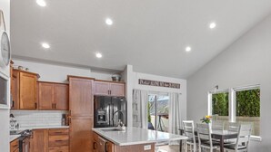 A kitchen with wooden cabinets, a stainless steel refrigerator, and a dining area with a table and chairs. - Spacious Modern Home—Fire Pit, Basketball Hoop, Pop-A-Shot & Kids' Toys for Family Fun! (Coeur D Alene)