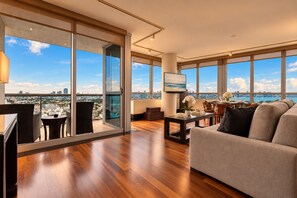 A spacious living area with a large window, a sofa, a dining table, and a balcony with a city view.