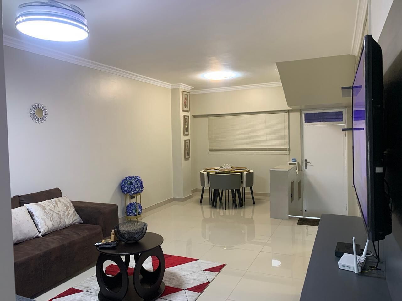 A modern living room with a sofa, coffee table, TV, and a dining area with chairs and a table.