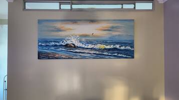 A painting of a seascape with waves and a bird, hung on a plain wall with a window above it.