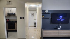 A modern living room with a flat-screen TV displaying the Golf Saudi logo, a kitchen with wooden cabinets and a red kettle, and a hallway leading to a bathroom. - A Two-bedroom Holiday Apartment, at the Heart of South Beach Durban (Durban)