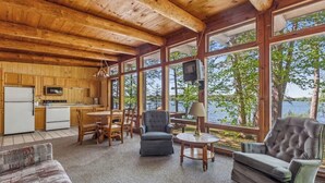 Une cabane comprenant une cuisine, un coin repas et un espace détente près d’une grande fenêtre donnant sur un lac.