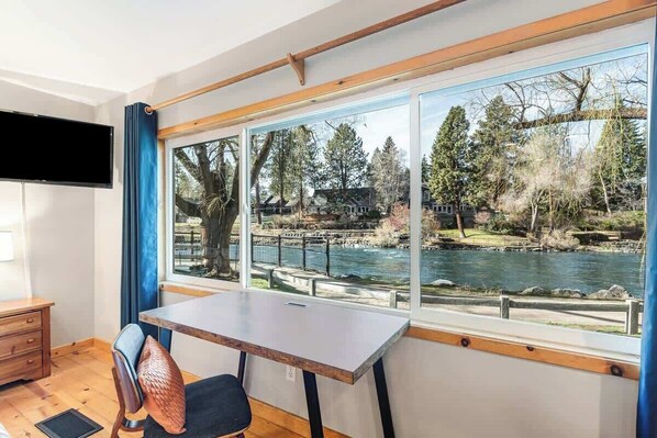 A room with a large window overlooking a river, a wooden desk, and a chair.