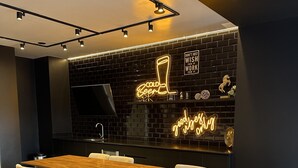 A modern dining area with a long wooden table and six chairs, a black brick wall with neon signs, and a black TV mounted on the wall. - Located in Zona 11, centrally-located,  family friendly, near airport, near mall (Guatemala City)