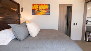 Un dormitorio con cama, almohadas, un cuadro, cabecera de madera y una puerta que da a una kitchenette.