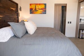 Une chambre à coucher comprenant un lit, des oreillers, un tableau, une tête de lit en bois et une porte donnant sur un coin cuisine.