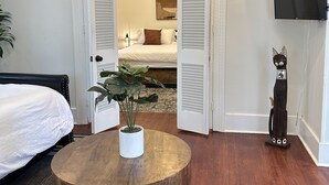 Una camera d'albergo con un letto, un tavolo rotondo in legno con una pianta in vaso, una TV montata a parete e una porta che dà su un'altra stanza.