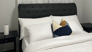 Un lit bien fait, avec une literie blanche et de nombreux oreillers, une tête de lit sombre recouverte de tissu et un rideau blanc.