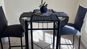 Un comedor con mesa de vidrio, dos sillas negras, una planta azul en maceta y un cuadro con botellas azules en la pared.