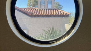 Una ventana redonda con vista a un edificio con tejado de tejas y una pequeña estructura tipo torre con ventanas arqueadas.