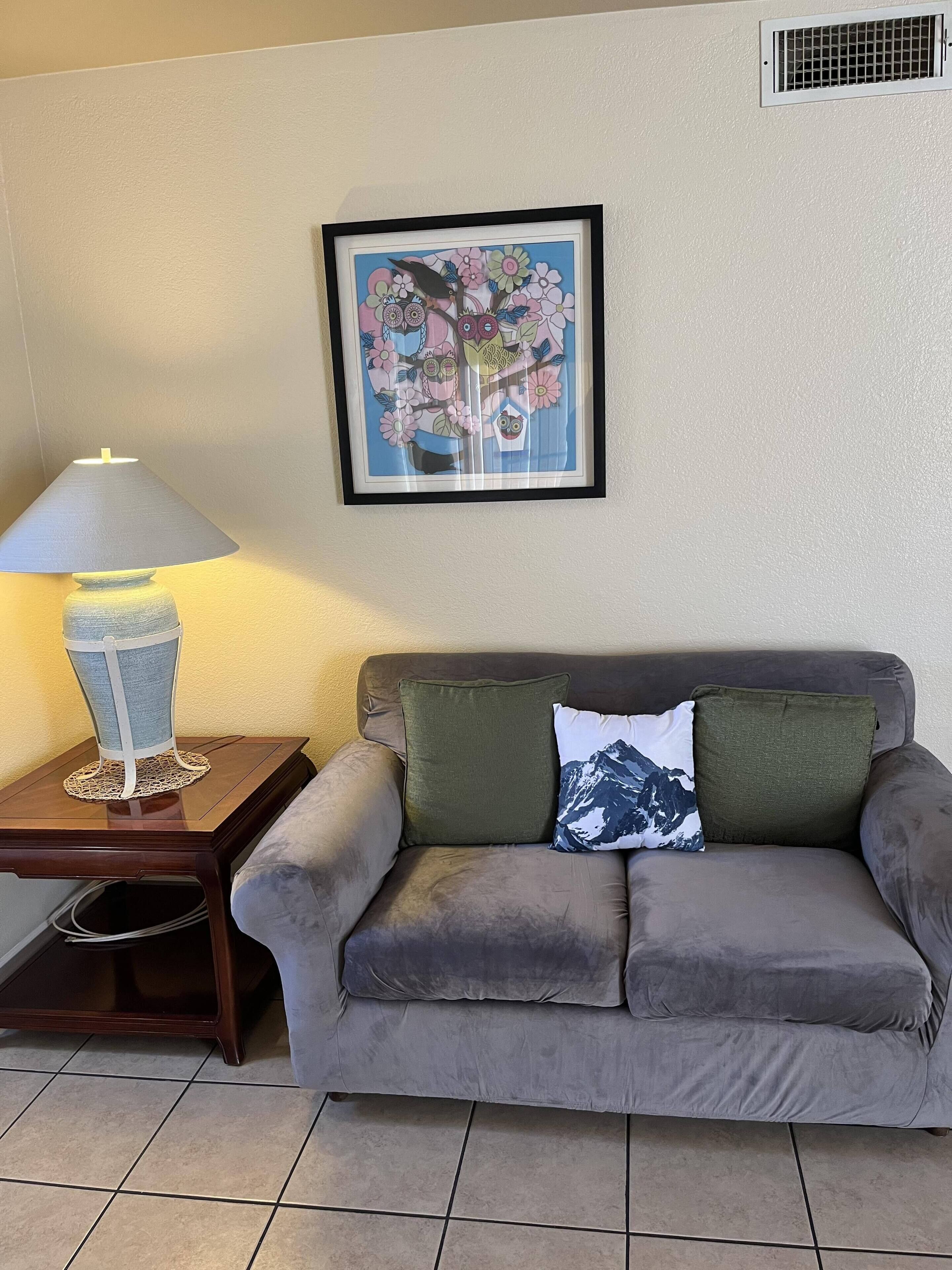 Una sala de estar con un sofá gris, una obra de arte floral enmarco, una lámpara de mesa y una pequeña mesa con un mantel de encaje.