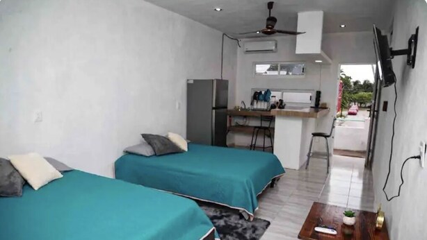 Una habitación compacta con dos camas individuales, una kitchenette pequeña, un ventilador de techo y un televisor montado en la pared.