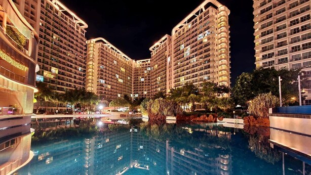 Vista nocturna de un hotel con varios edificios altos, una piscina central y vegetación circundante.