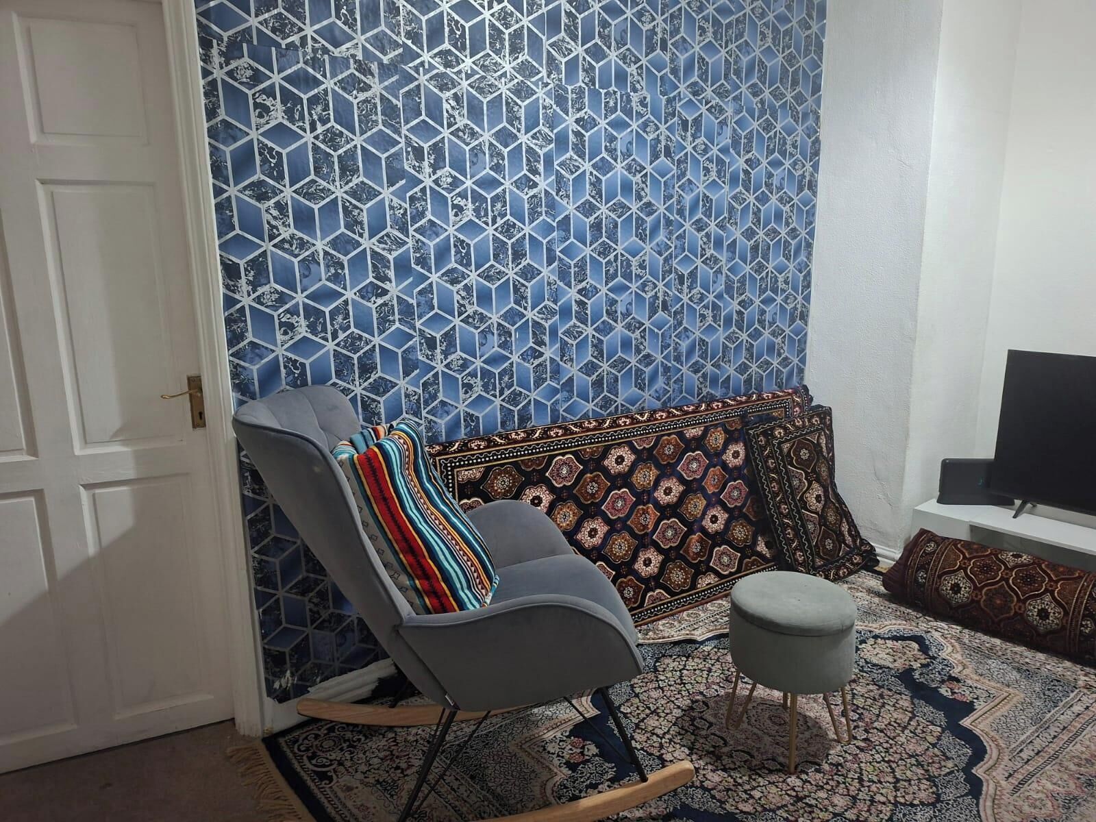 A room with a patterned wall, a grey rocking chair, a small ottoman, and a television on a stand.