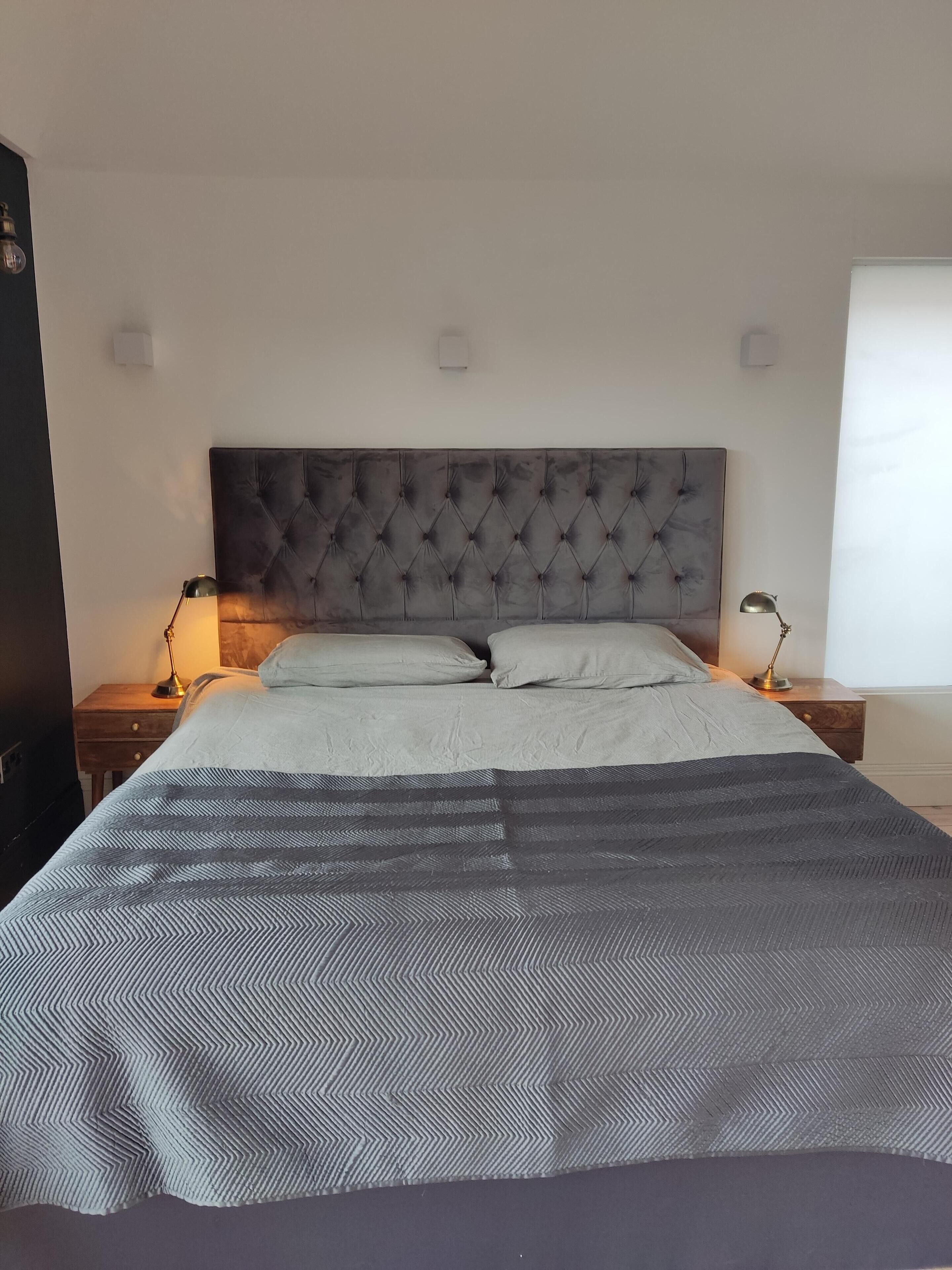 Ein ordentlich bezogenes Bett mit einer karplierten Kopfteil, zwei Nachttischlampen und einem Fenster mit Jalousien.