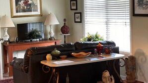 A living room with a black leather sofa, a wooden coffee table, a television on a wooden stand, and a window with blinds.