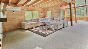 Ein geräumiges Loft mit einer großen Eckcouch, Holzbalken und großen Fenstern mit Blick auf den Wald.