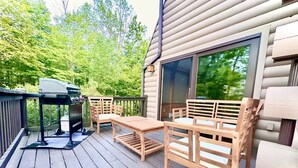 Eine Terrasse mit Holztisch und -stühlen, einem Grill und einem großen Fenster mit Blick auf ein bewaldetes Gebiet.