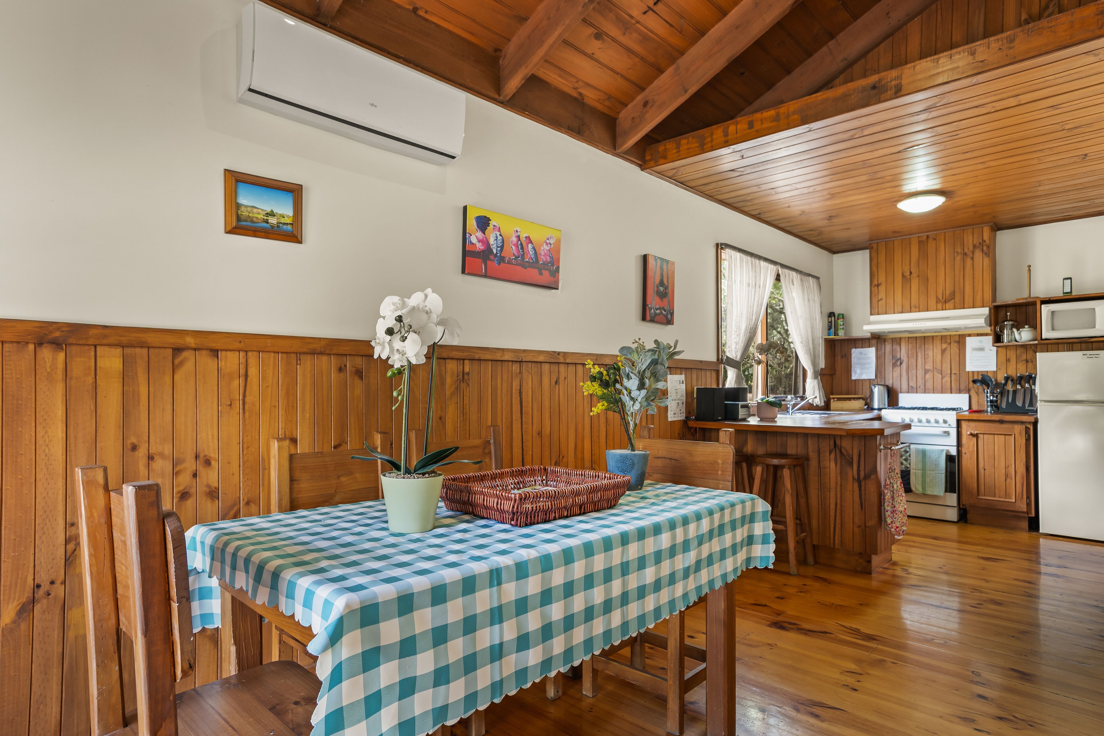 A wooden interior with a dining table covered by a checkered tablecloth, wooden chairs, a kitchen area with wooden cabinets, and a microwave.
