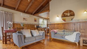 A cozy living room with wooden floors and furniture, a dining area with a checkered tablecloth, and a kitchen in the background.