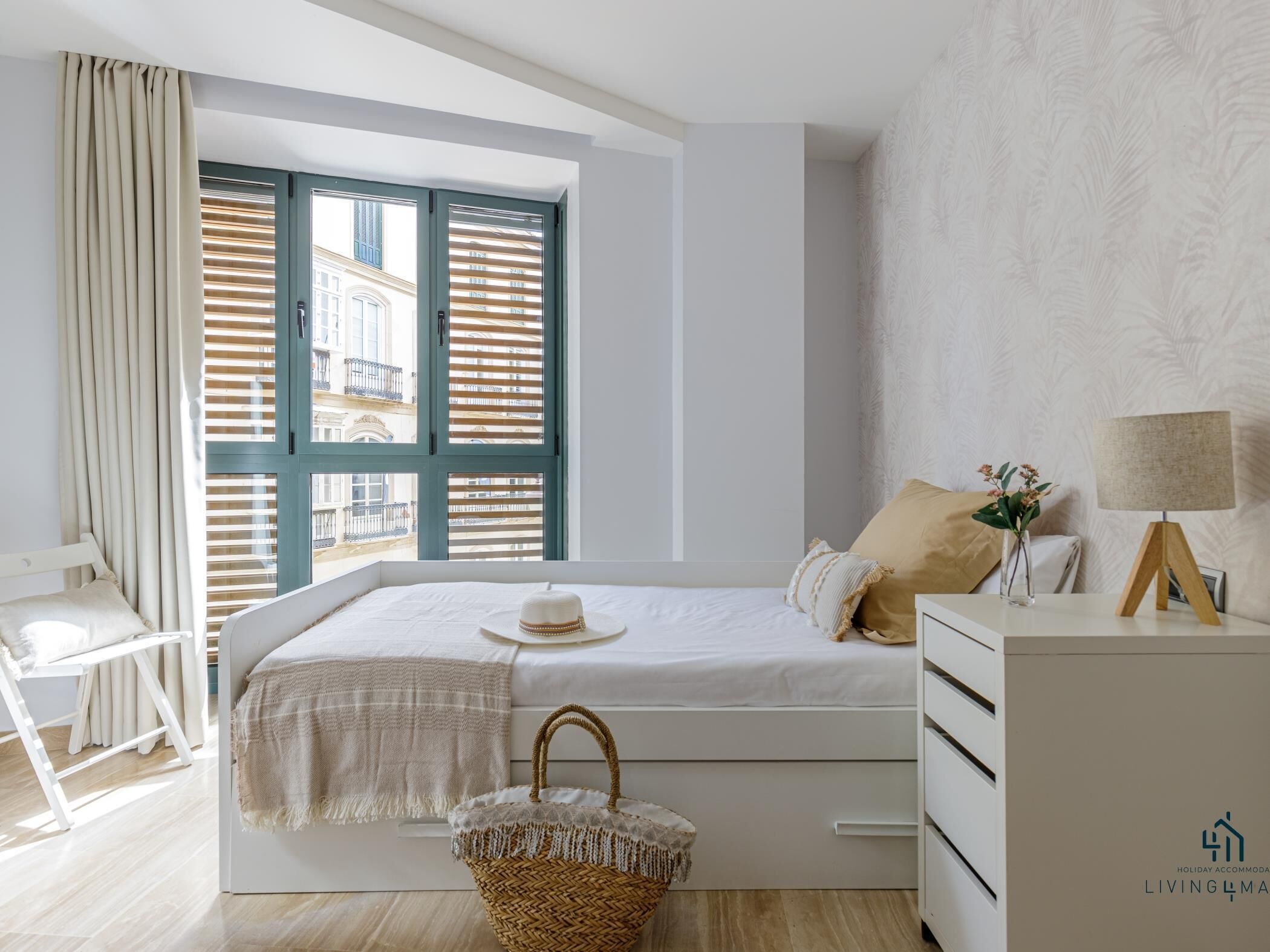 Un dormitorio con cama, mesita de noche con lámpara, una silla, ventana con persianas y vistas a edificios.