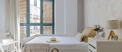Un dormitorio con cama, mesita de noche con lámpara, una silla, ventana con persianas y vistas a edificios.