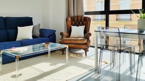 Een woonkamer met een blauwe bank, een leren fauteuil, een glazen salontafel, en een eethoek met een glazen tafel en stoelen.