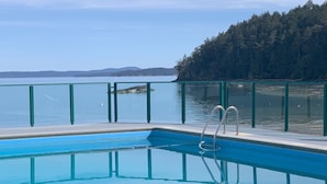Una piscina con una escalera metálica, rodeada por una barandilla verde, con vista a una masa de agua tranquila y una ladera boscosa.