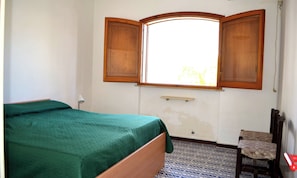 A room with a bed, a chair, a window with wooden shutters, and a small shelf.