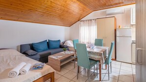 A compact room with a wooden ceiling, a sofa, a dining table with chairs, a bed with a rolled-up towel, and a kitchen area with a refrigerator. - Studio Apartment Iva Rijeka Suburbs, Rijeka, Croatia (Rijeka)