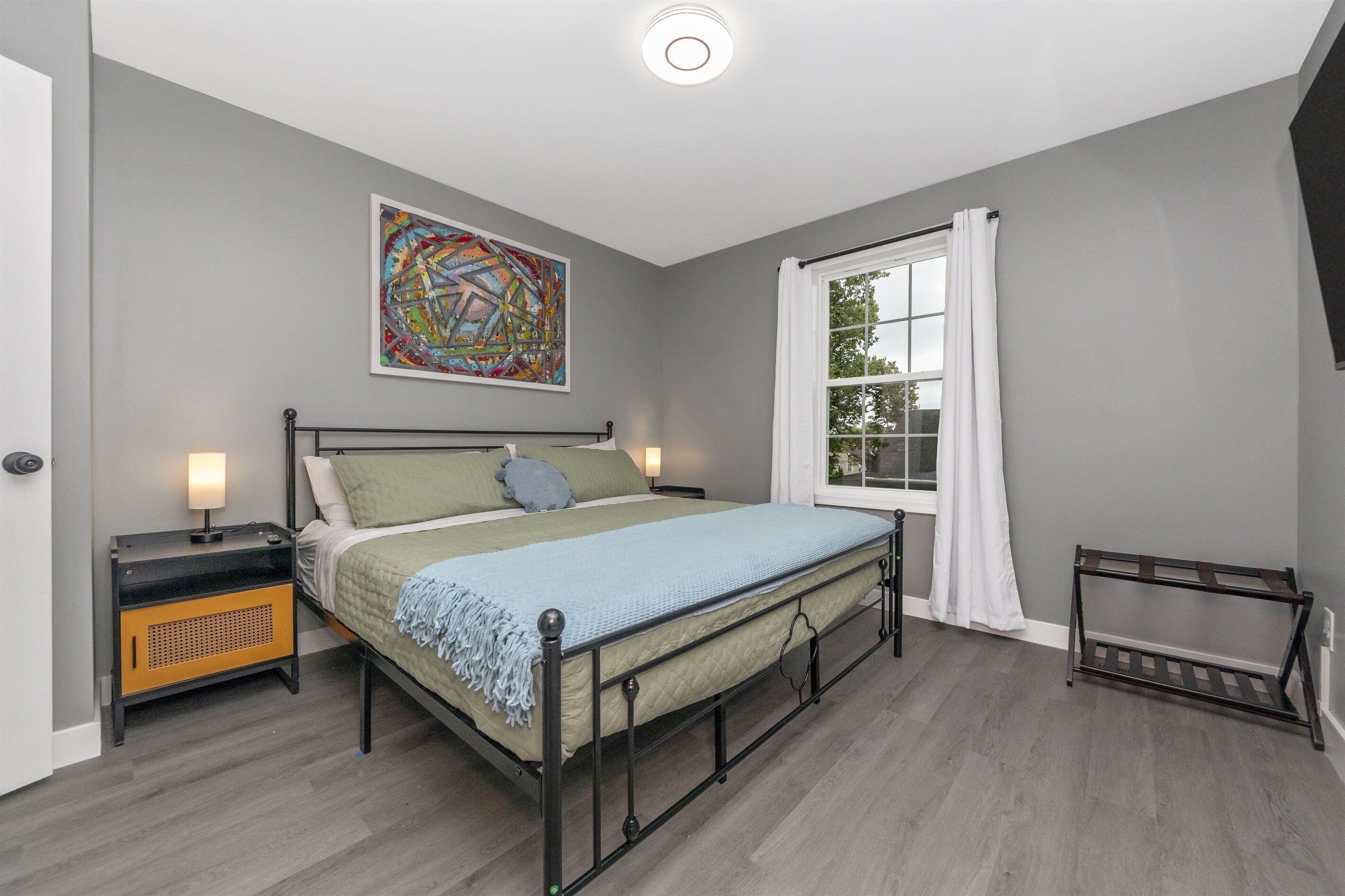 Une chambre à coucher comprenant un lit, une table de chevet, une fenêtre avec des rideaux et une œuvre d’art accrochée au mur.
