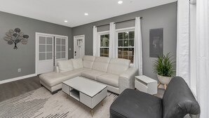 En moderne stue med en hjørnesofa, et sofabord og en grå sofa. Der er en potteplante og en hvid vægdekoration.