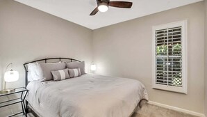 Een slaapkamer met een bed, een plafondventilator, een nachtkastje met een lamp en een raam met jaloezieën.