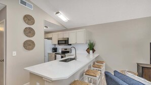 Een compacte keuken met witte kastjes, een magnetron en een spoelbak. In de aangrenzende woonruimte staan drie geweven krukken en een blauwe bank.