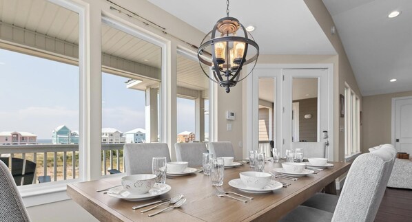 Un coin repas avec une longue table en bois, dressée pour quatre personnes, de grandes fenêtres et une vue sur la plage.