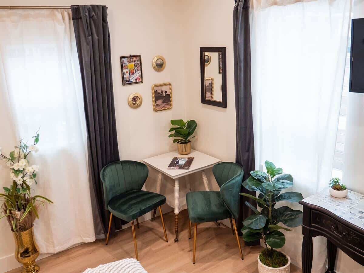 A small, cozy room with a white table, two green chairs, a potted plant, and a mirror on the wall.