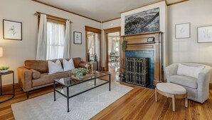 A living room with a brown sofa, a white armchair, a glass coffee table, and a fireplace with a framed picture above it.