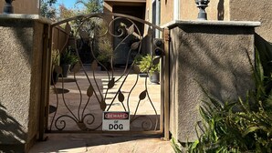 Une entrée fermée par un portail, agrémentée d’un panneau d’avertissement « Attention, chien », un portail en fer forgé et un escalier en pierre menant à un bâtiment doté d’un toit en tuiles et de murs en stuc.