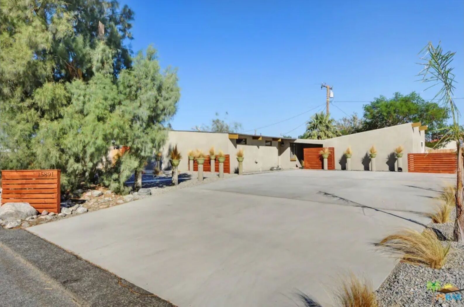 Une allée spacieuse mène à une maison moderne dotée d’un grand garage, le tout entouré d’un jardin bien entretenu et de grands arbres.