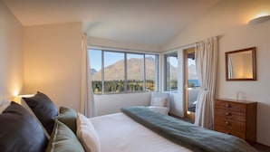 Une chambre avec une grande fenêtre donnant sur les montagnes, un lit recouvert d’un plaid vert, une commode en bois et un miroir au mur.