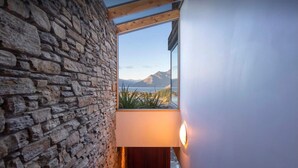Une pièce avec un mur en pierre, une fenêtre donnant sur les montagnes et l’eau, et un luminaire fixé au mur.