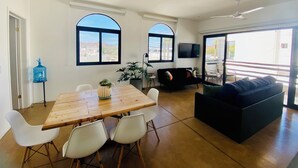 Un amplio salón con una mesa de comedor de madera, sillas blancas, un sofá negro y grandes ventanales que ofrecen vistas de la ciudad.