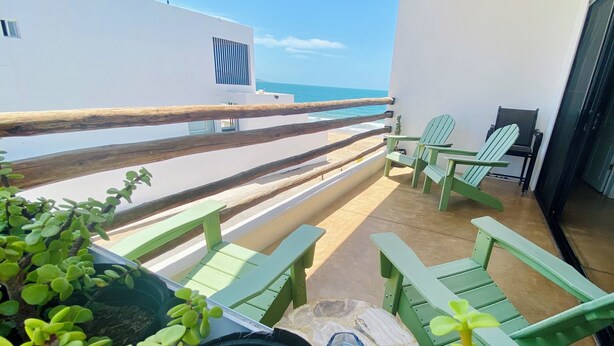 Un balcón con vista al mar, sillas verdes, una mesita y una planta en maceta.