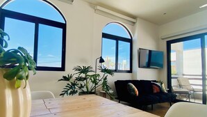 Una sala de estar moderna con una mesa de madera, dos amplias ventanas de arco y un sofá negro, con un televisor montado en la pared.