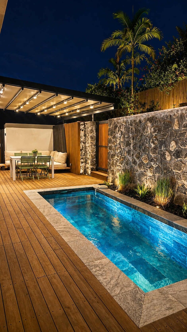A swimming pool with a wooden deck, a stone wall, and a covered seating area.