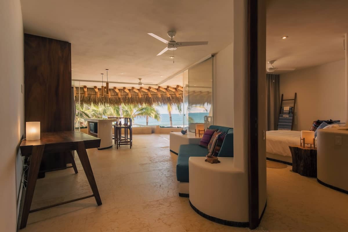 Een moderne hotelkamer met een groot bed, een zithoek met uitzicht op de oceaan, een eethoek en een plafondventilator.