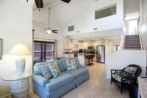 A spacious living area with a sofa, a glass coffee table, a chair, a lamp, and a kitchen in the background.
